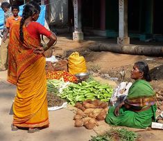 two women are sitting on the ground in front of vegetables and fruit, one is looking at another woman