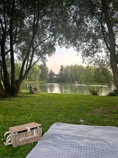 a picnic blanket and basket sitting on the grass next to a lake with trees in the background