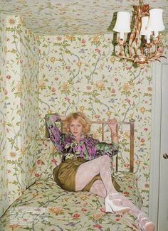 a woman sitting on top of a bed in a room with floral wallpaper and chandelier