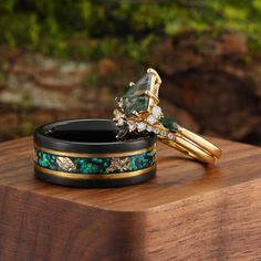 two gold rings with green and blue stones on them sitting on a wooden table next to some rocks