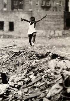 a black and white photo of a young boy jumping in the air over rubble covered ground