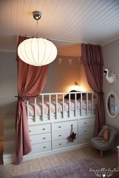 a bedroom with pink curtains and white dressers in front of the bed is lit by a light fixture