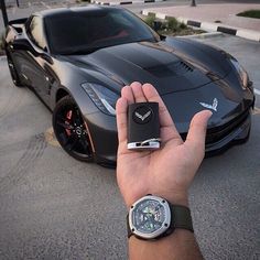 a person holding a cell phone in front of a black sports car on the street