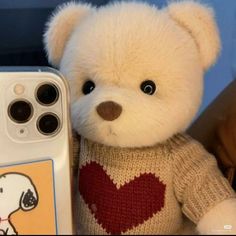 a white teddy bear with a red heart on it's sweater next to an iphone