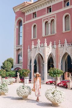 a woman standing in front of a large pink building with white flowers on the ground