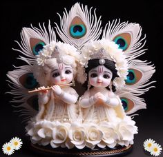 two figurines sitting on top of a table next to white flowers and peacock feathers