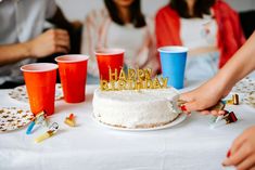 people are sitting at a table with a birthday cake and cups in front of them