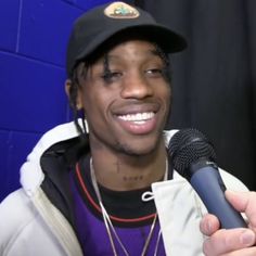 a man holding a microphone in front of a blue wall and wearing a black hat