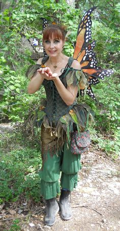 a woman dressed as a fairy holding a butterfly on her shoulder and standing in the woods