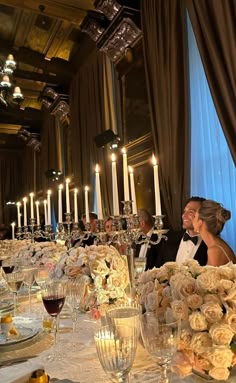 a man and woman sitting at a table with flowers in front of them, surrounded by candles