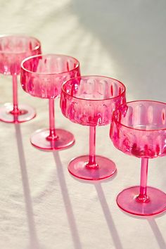 three pink wine glasses sitting on top of a white table covered in light shining from the window