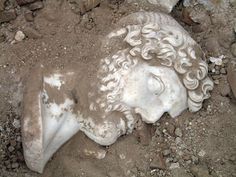 an old statue laying in the dirt on top of some rocks and gravel with it's head covered by hair