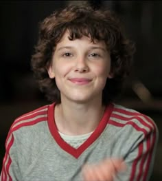 a young boy with curly hair is smiling and holding something in his hand while wearing a red and gray shirt