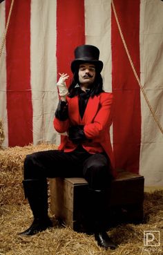 a man in a top hat and red jacket sitting on a box with his hands up