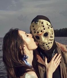 a woman kissing a man wearing a mask in front of the water with trees behind her