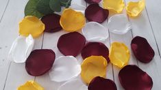 several different colors of petals on a white wooden surface with green leaves and flowers in the background