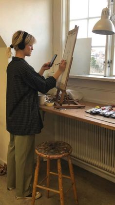 a woman standing in front of a easel painting on an easel next to a window