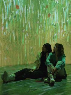 two women sitting on the ground in front of a painting with grass growing behind them