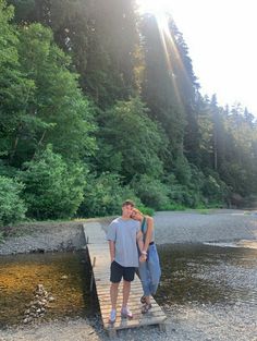 a man and woman standing on a wooden bridge over a river with trees in the background