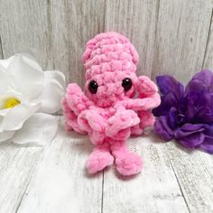 a pink crocheted stuffed animal sitting next to two white and purple flowers on a wooden surface