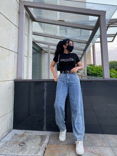 a woman in black shirt and jeans standing next to a wall wearing a face mask