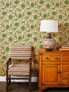 a chair sitting next to a dresser with a lamp on top of it and a wall paper behind it