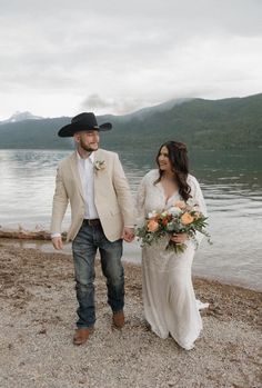 a bride and groom walking on the shore of a lake holding hands with each other