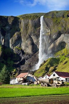 a large waterfall towering over a lush green field next to a rural area with farm buildings