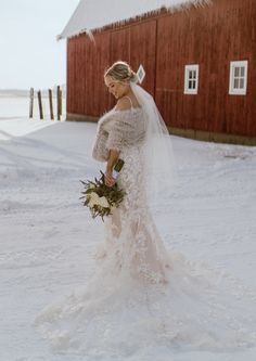 a woman in a wedding dress standing in the snow next to a red barn and holding a bouquet