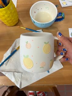 a person holding a cup on top of a wooden table next to a bowl and pencils