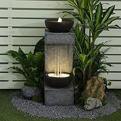 a water fountain with lit candles in it next to some plants and rocks on the ground