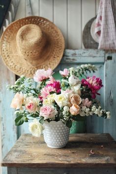 a white vase filled with lots of flowers on top of a wooden table next to a hat