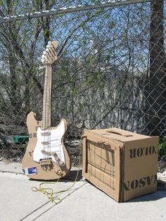 an electric guitar sitting on the ground next to a cardboard box and chain link fence