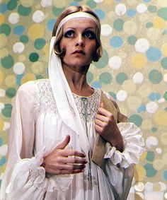 a woman wearing a white dress and head scarf with her hands on her chest, standing in front of a colorful background