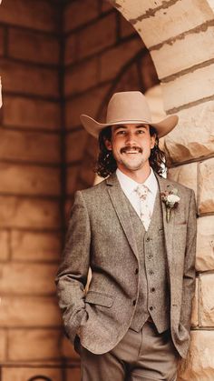 a man in a suit and hat standing by a stone wall with his hands on his hips