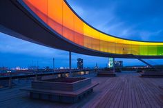 an empty wooden deck with benches under a rainbow - colored awning at dusk in the city