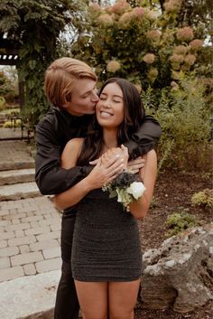 a man and woman hugging each other in front of some bushes with flowers on them