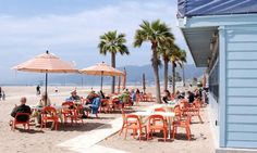 people are sitting at tables on the beach under umbrellas and chairs with palm trees in the background