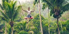 a man on a swing in the middle of some trees and palm trees with his arms outstretched