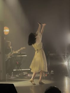 a woman standing on top of a stage holding her arms up in the air while wearing a yellow dress