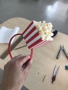 a hand holding a red and white striped popcorn box with scissors on the table next to it