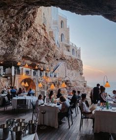 people are sitting at tables in front of an ocean side restaurant with large cliffs behind them