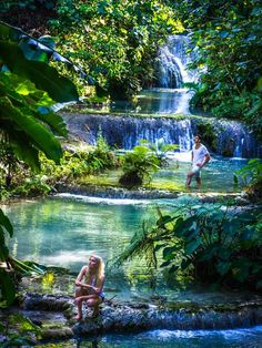 two people sitting on rocks in front of a small waterfall with water running down it