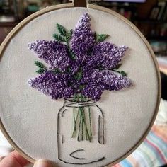 a person holding up a small embroidery project with flowers in a mason jar on it