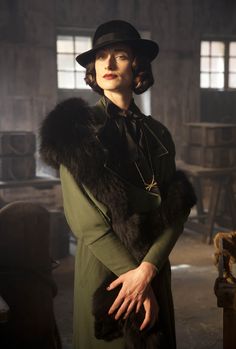 a woman in a green coat and black hat is posing for the camera with her hands on her hips