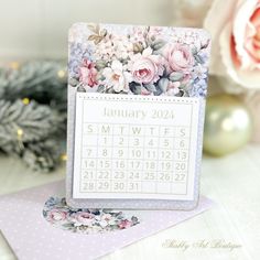 a calendar card with flowers on it sitting next to some christmas trees and ornaments in the background