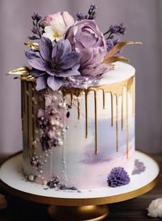 a white cake with purple flowers and gold drips on top, sitting on a wooden table