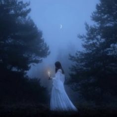 a woman in a white dress is holding a candle and looking into the distance with trees behind her