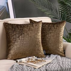 two gold pillows on a white couch next to a book and some silver beads in front of a window