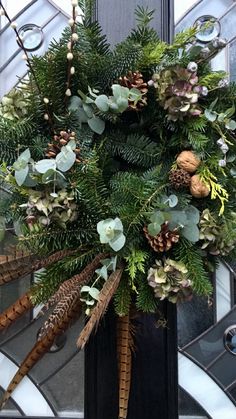 a wreath with pine cones and evergreens is hanging on the front door, surrounded by greenery